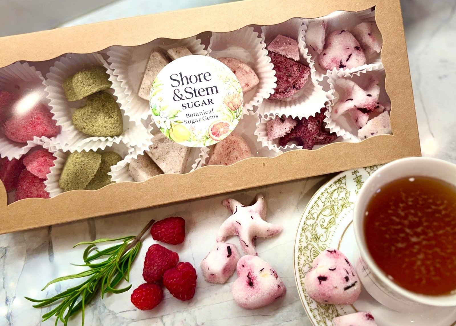 Box of Shore & Stem Beverage Gems, a teacup of tea, and raspberries and Rosemary on a marble surface.